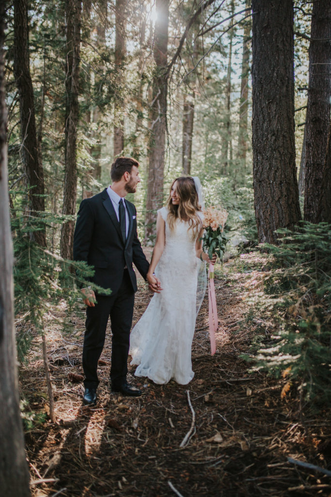 bride and groom walking through the forest in the Smoky Mountains holding hands. Bride is in a lace dress and had a romantic pink bouquet of flowers. Located in Knoxville Tennessee