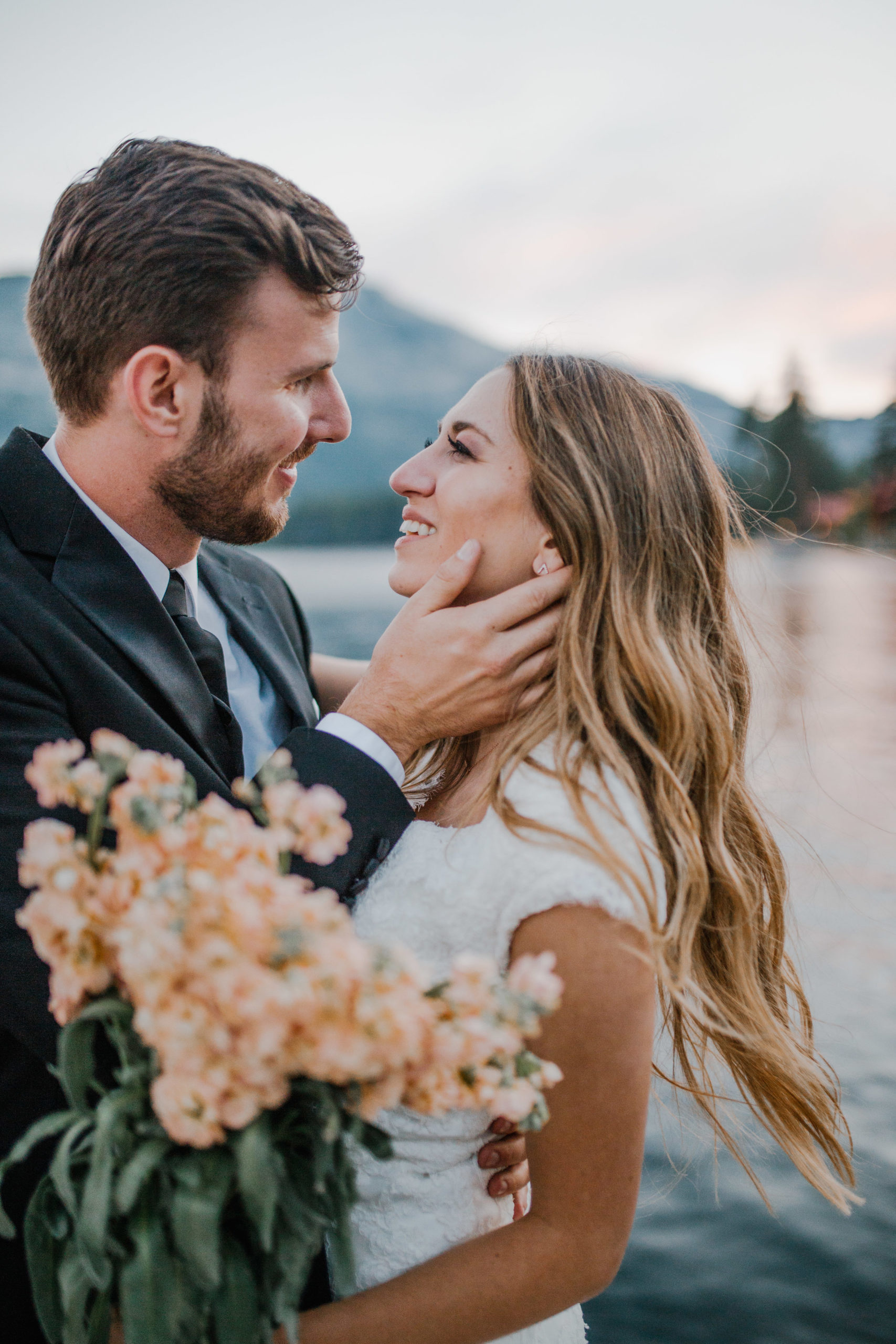 bride and groom standing on a pier on a lake in Nashville Tennessee looking at each other while the groom hold the brides face and she smiles while holding a peach bouquet at sunset