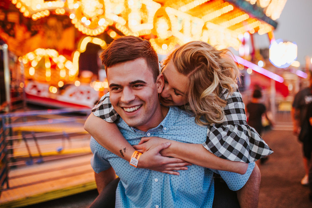 engagement session at a carnival with the woman on the mans back for a piggy back ride in front of a carousel 