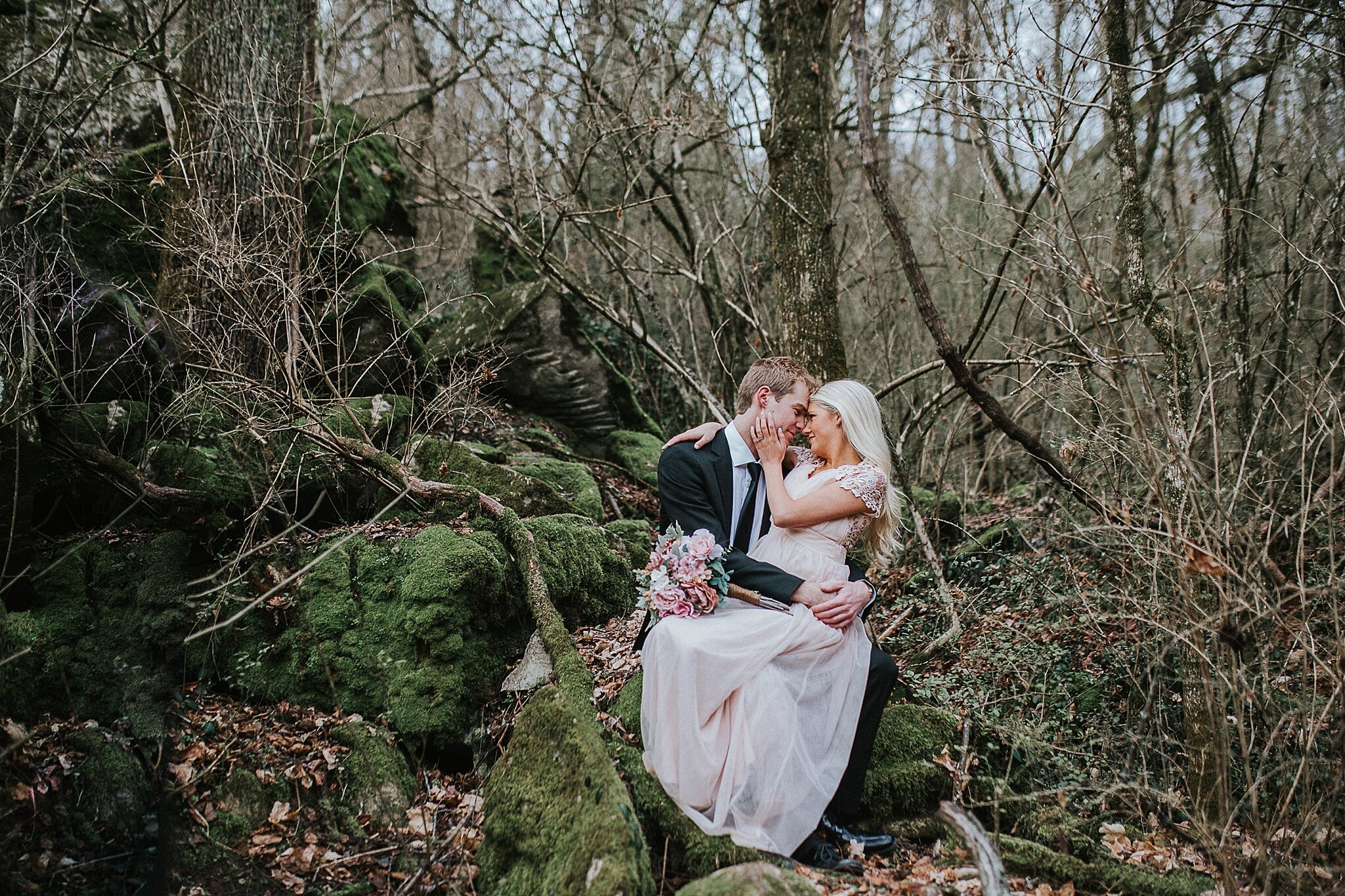 bride sitting on grooms lap with her arms around his neck pullin ghim closer to her face as he holds her around the waist as they sit oon a mossy stone in the woods