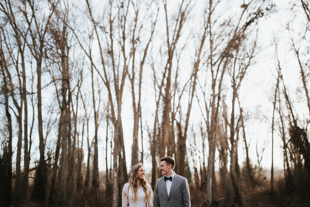 bride and groom standing infront of bare fall trees looking at eachother with love