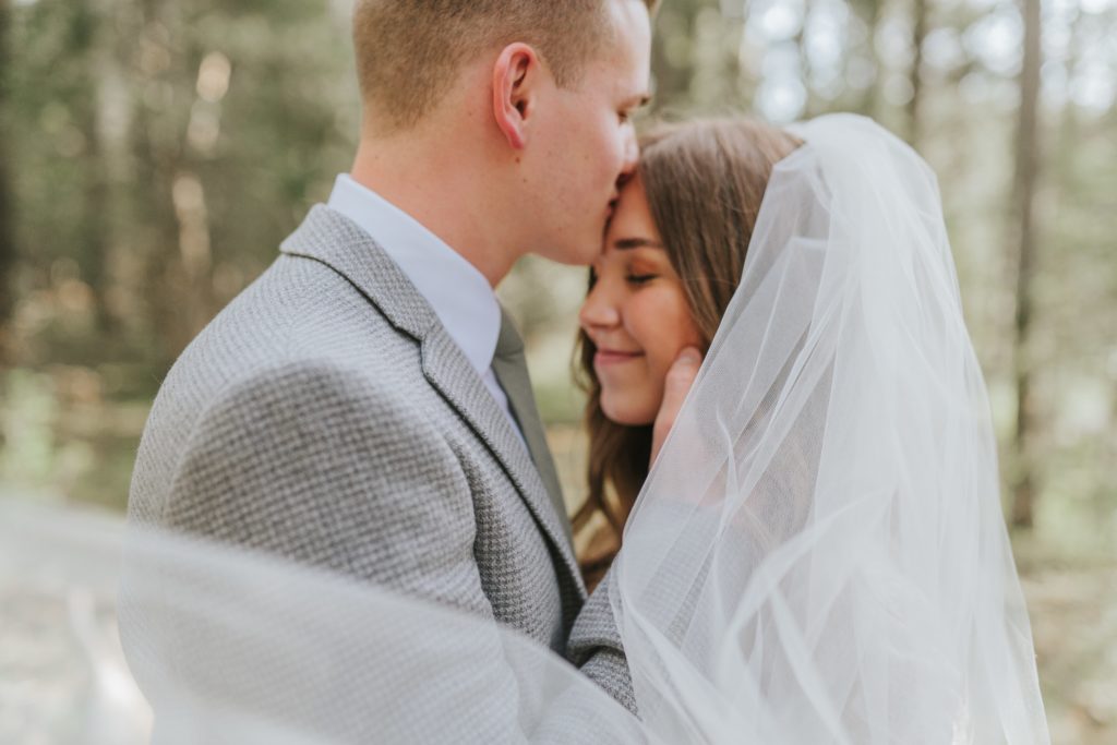 groom kissing bride's forehead during forest bridals while veil blows in the wind
