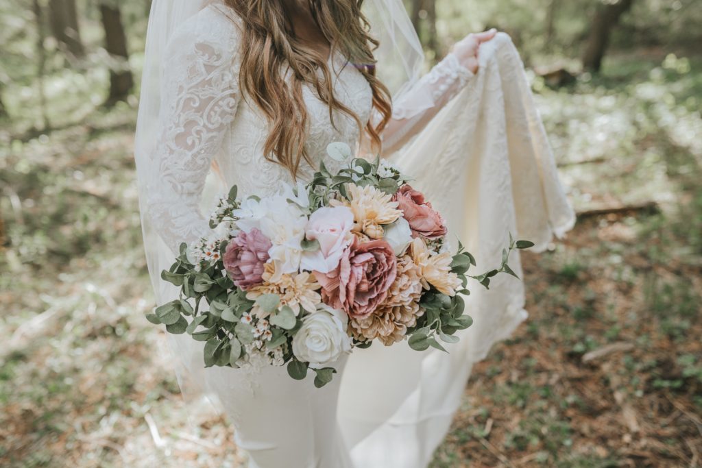 Bride standing in a forest in her wedding dress while holding her bouquet of white, pink, and yellow flowers
