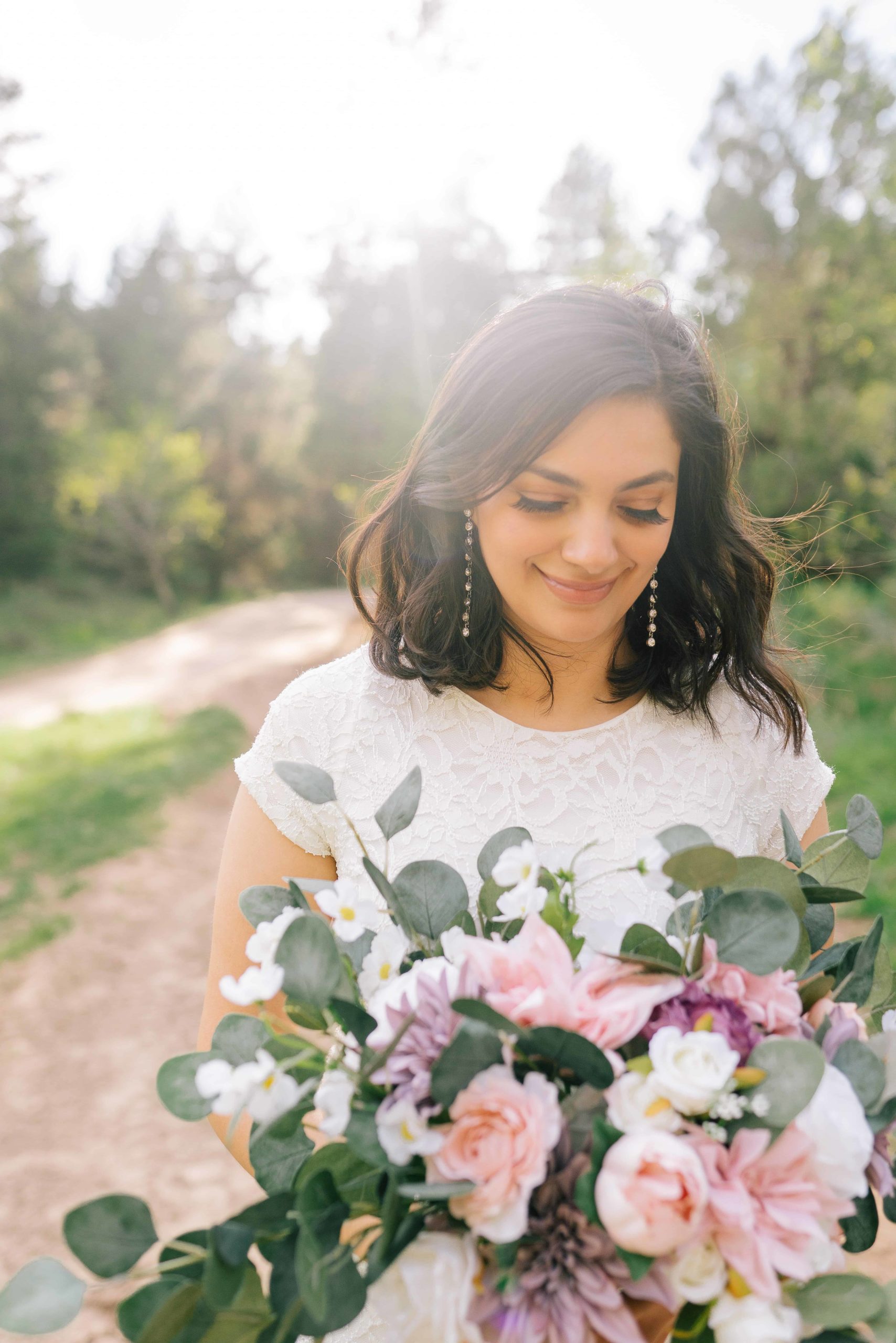stunning Latina bride holding her wedding bouquet of soft pinks and purples smiling as she looks down with her eyes and she sun shine from behind her