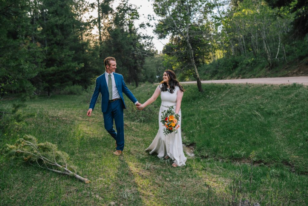 plana smoky mountain wedding in 6 months man in blue suit holding hands with beautiful bride in a lace summer gown while walking through the woods on their wedding day