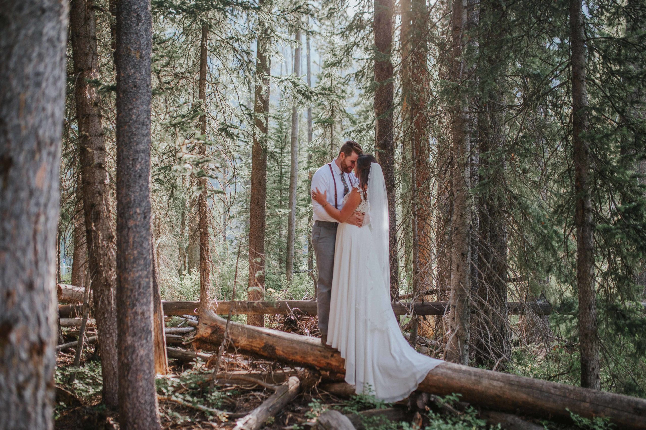 bride and groom standing on a log in the middle of the woods holding hands and looking at each other