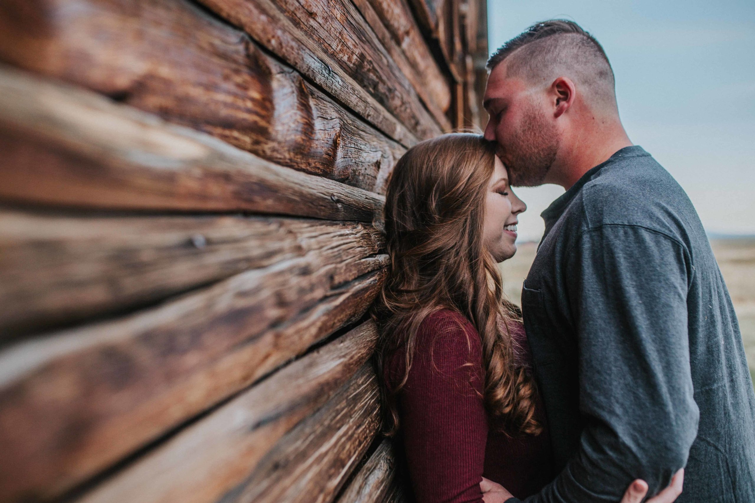 husband kissing wife on the forehead against the cabin in the winter