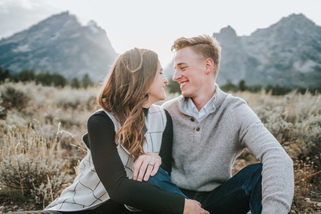 boyfriend and girlfriend sitting with each other for an engagement session in the Smokey Mountains. Fiancés are staring at each other while holding hands romantically. Mountain range in the background with bright light shining through.