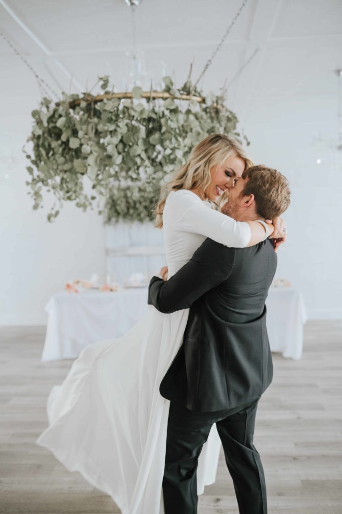 wedding bride and groom in a gorgeous reception hall. groom twirling bride in the air. White room with eucalyptus leaf chandelier
