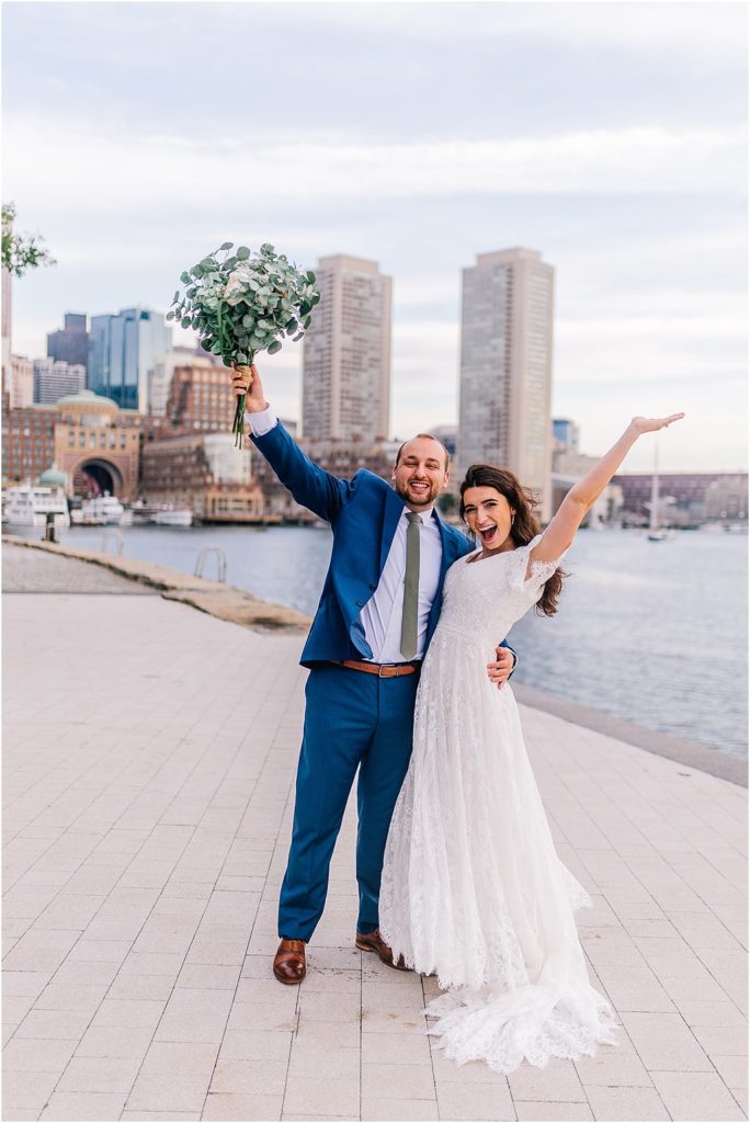 bride and groom in front of the water holding bouquet with hands up in celebration