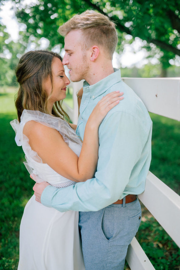 groom is holding his bride near a white fence and they are smiling at each other at Marblegate Farm in Knoxville Tennessee. Green summer grass is surrounding the wedding couple.