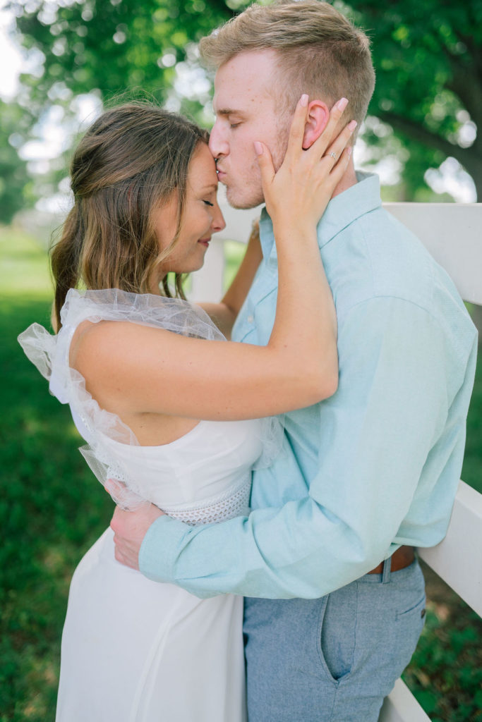 husband is kissing his wife on the forehead as she holds his jaw and he holds her waist. The wedding couple is leaning on a white fence at Marblegate Farm in Knoxville Tennessee