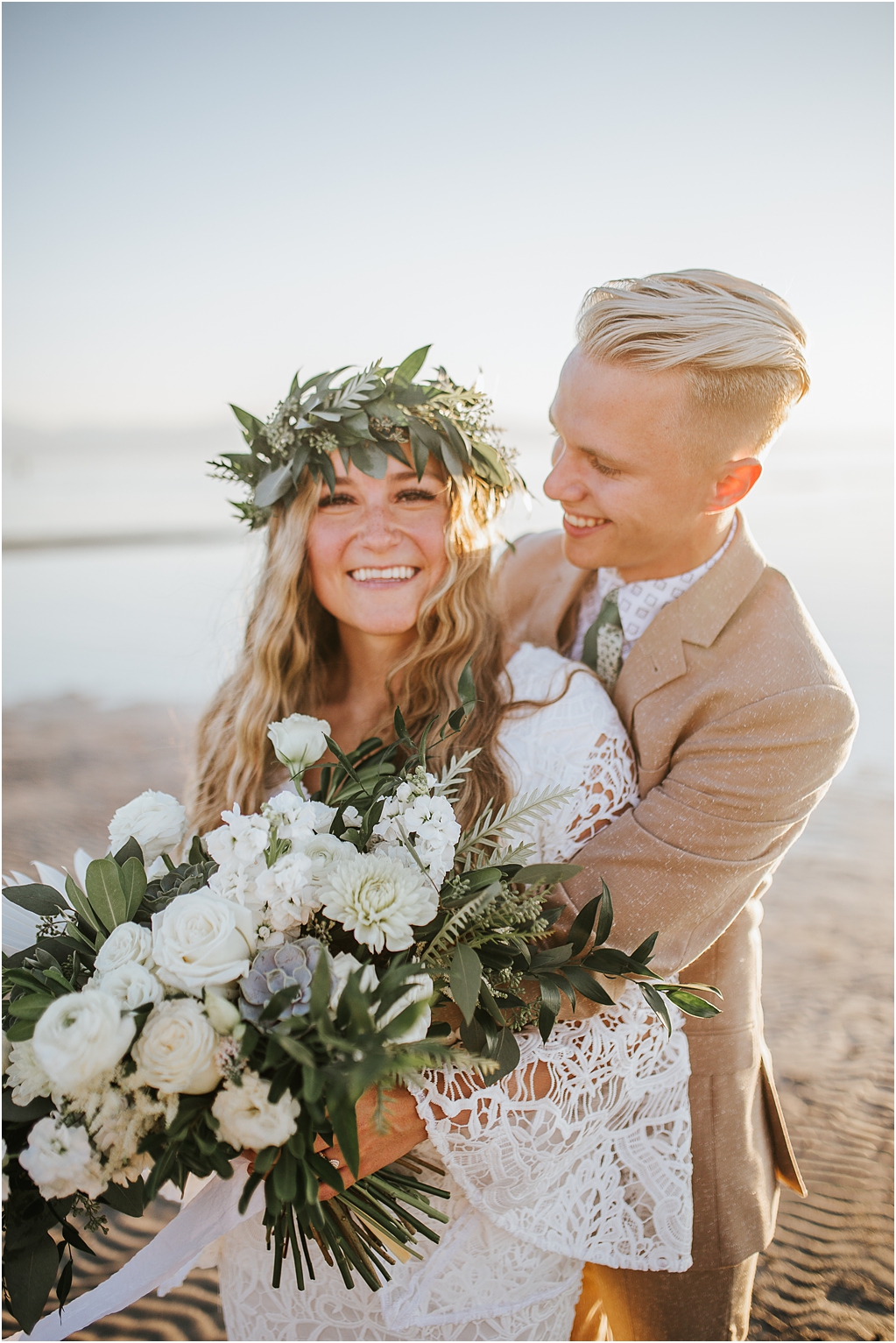 bride and groom for their boho bridals with groom hugging bride from behind and smiling at her as she looks at the camera and smiles, bride wearing a flower crown and a lace bohemian wedding dress with a large white rose bouquet