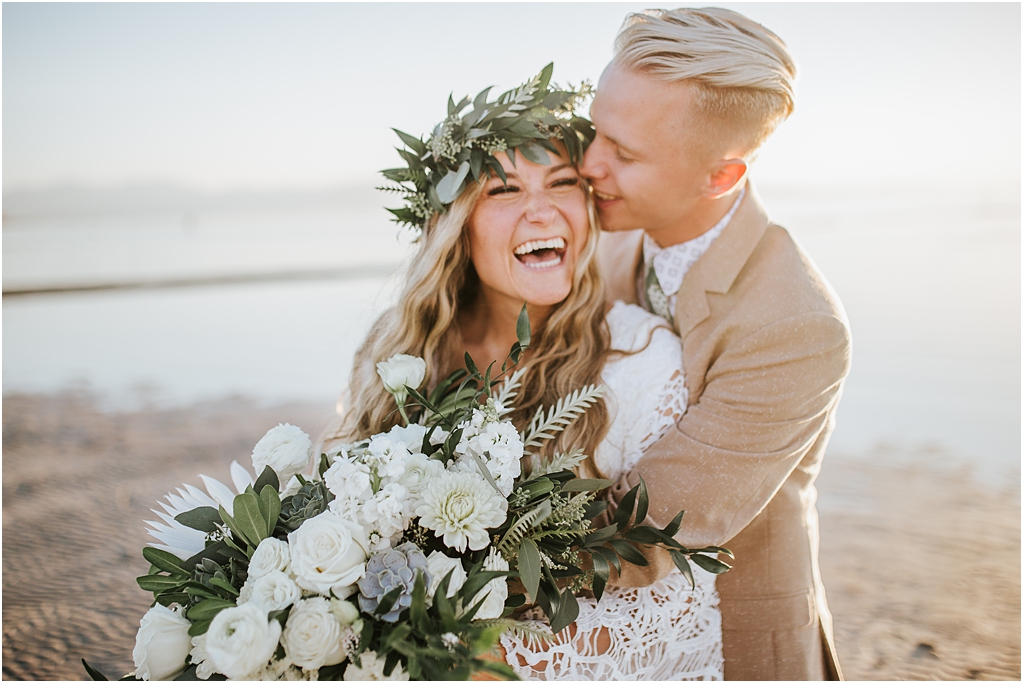 Bride and groom at the outerbanks in North Carolina on the beach laughing together and smiling as the sun goes down behind them. Bride is in a lace white gown and holding all white rose bouquet
