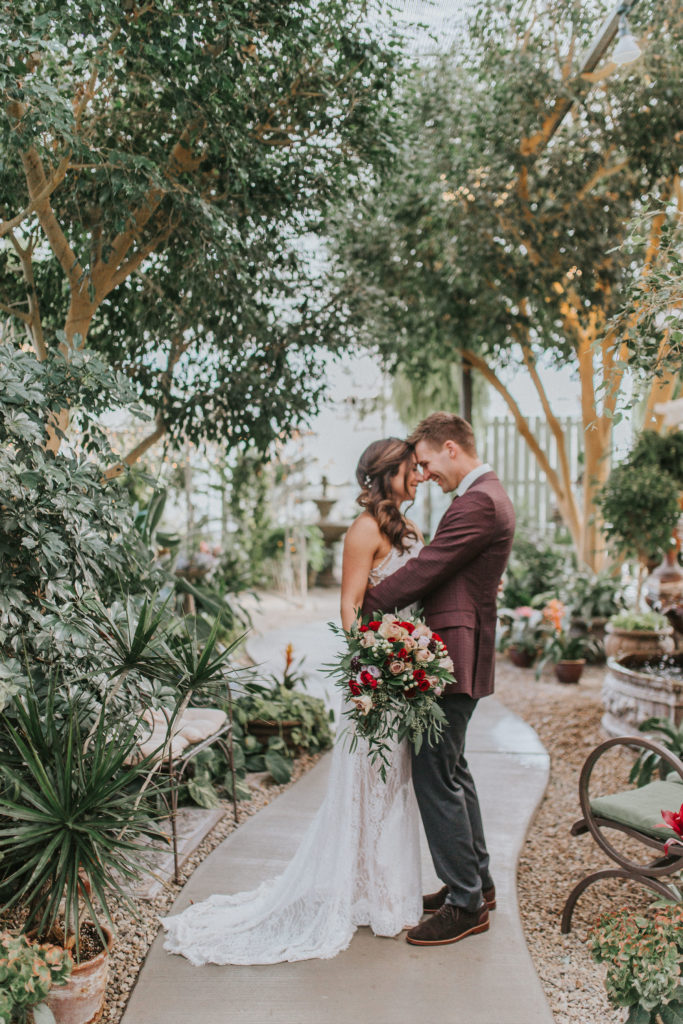 wedding couple embracing each other surrounded by flowers and plants, holding a red rose bouquet 