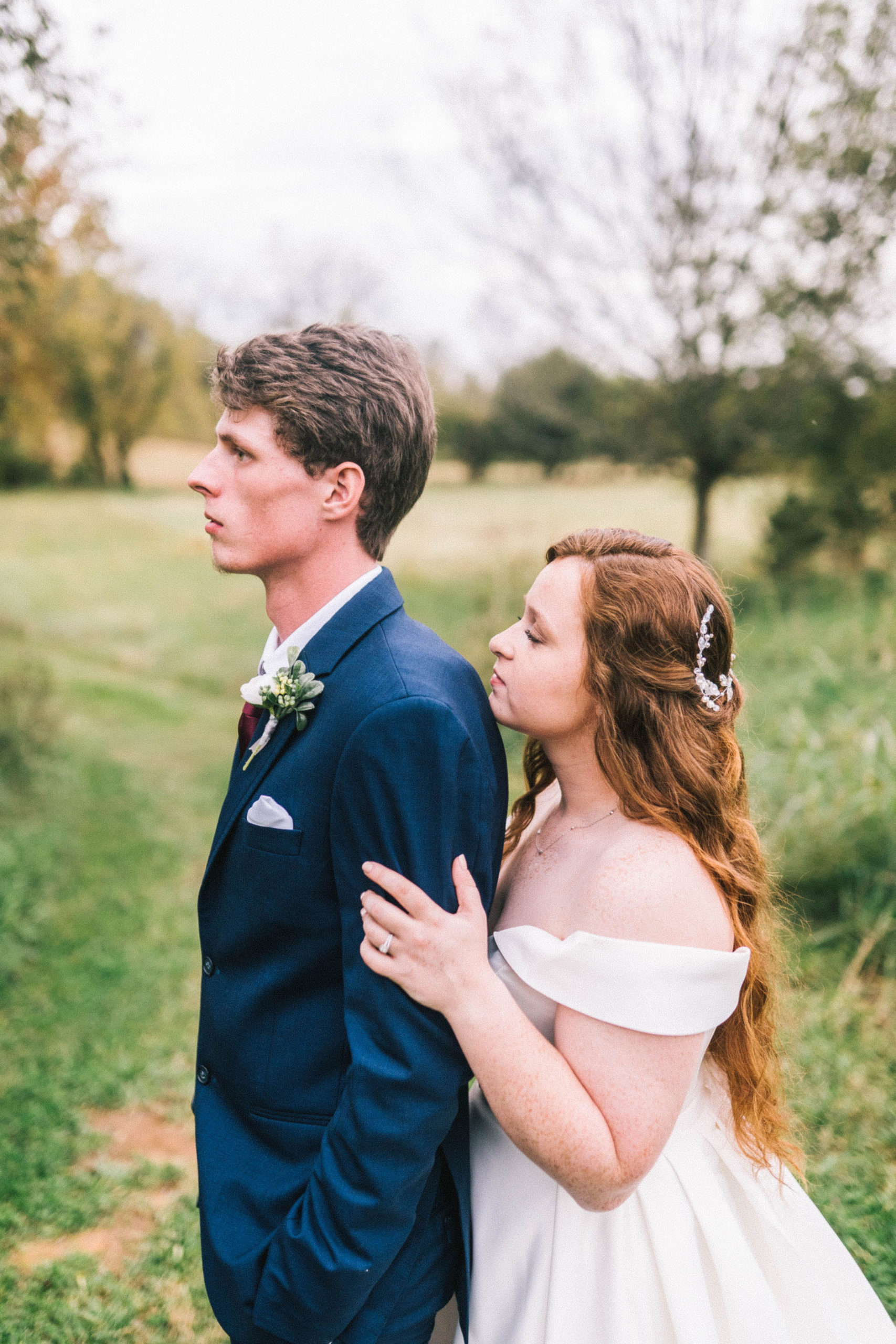 Bride and groom at butler farms in Knoxville Tennessee on their wedding day. Groom in a blue suit and facing away from his bride. Bride holding her husband and looking over his sholder. Profile picture of husband and wife