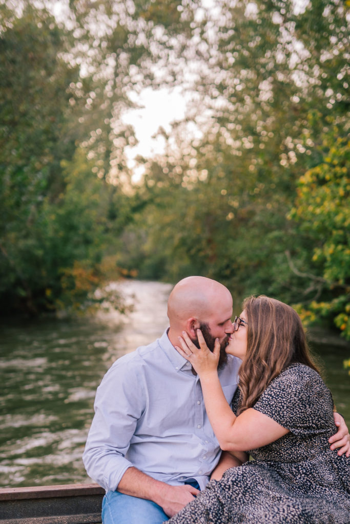 sitting on a bridge and kissing each other at seven islands birding pond in the fall, engagement session