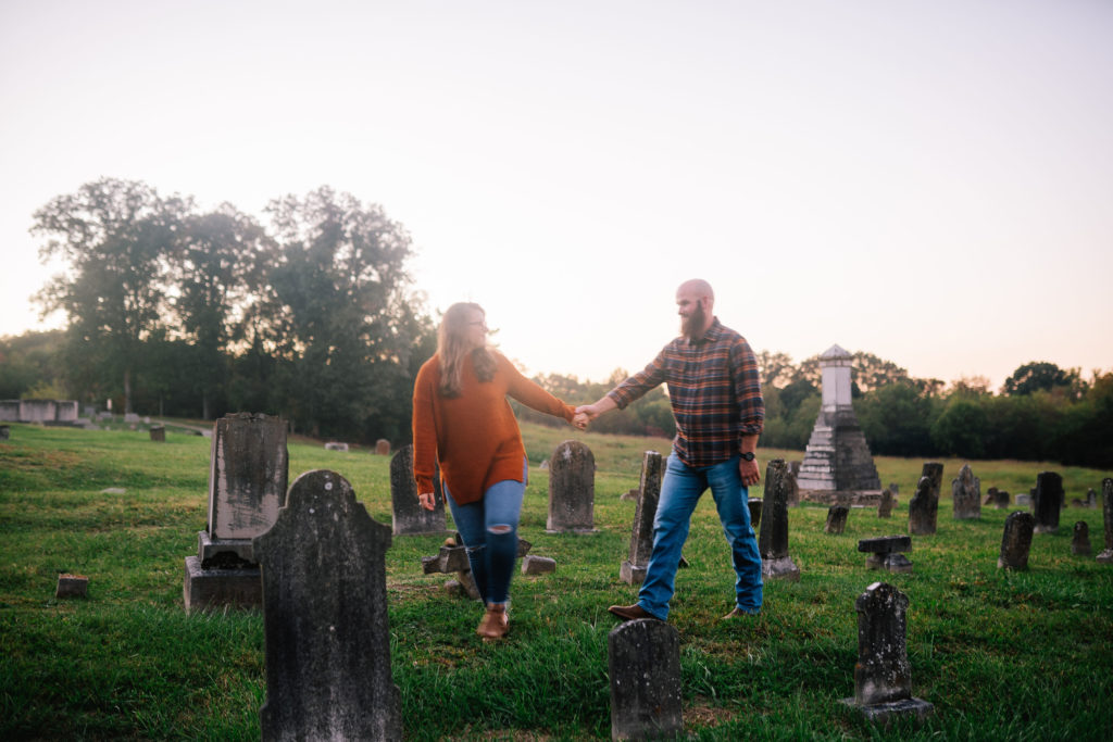 engagement shoot in a grave yard. The couple is holding hands and the woman is leading the man through the graveyard at seven islands birding pond