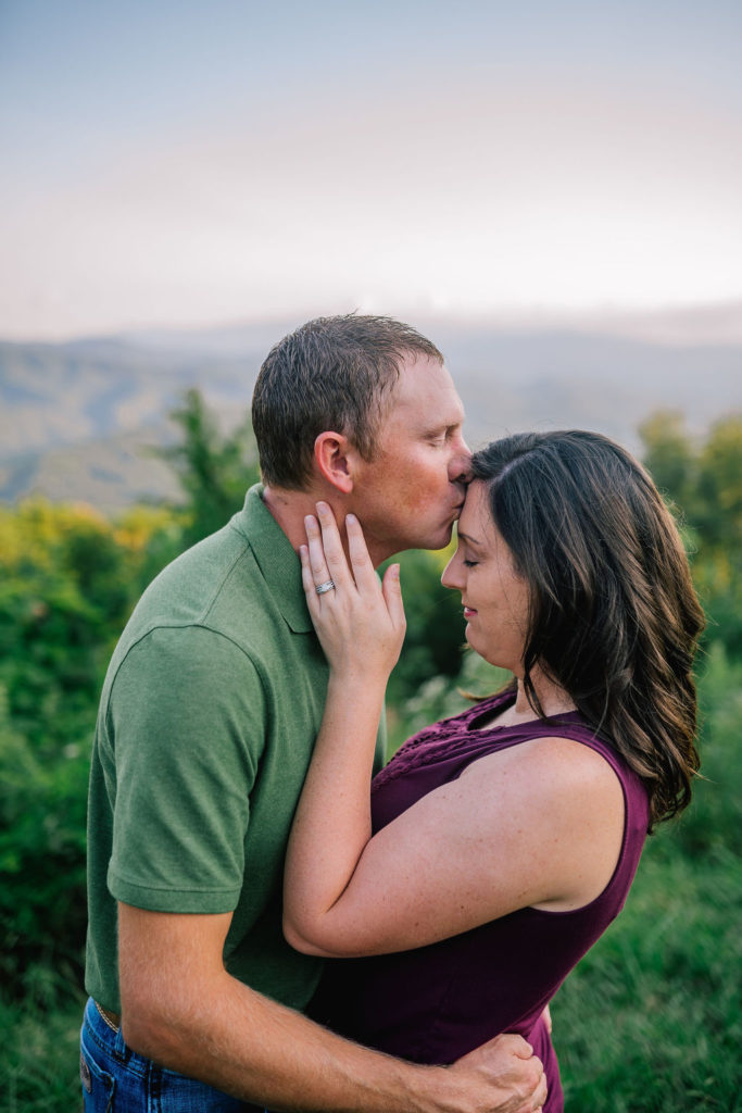 married couple in the Smokey Mountains, husband embracing his wife around the waist and kissing her forehead, wife holding husbands face with her eyes closed