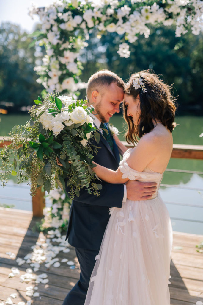 groom holding his bride on a southern river under a wedding arch. bride is wearing and elegant white wedding gown and holding a white bouquet. Bride and groom have their foreheads together and smiling on their wedding day.