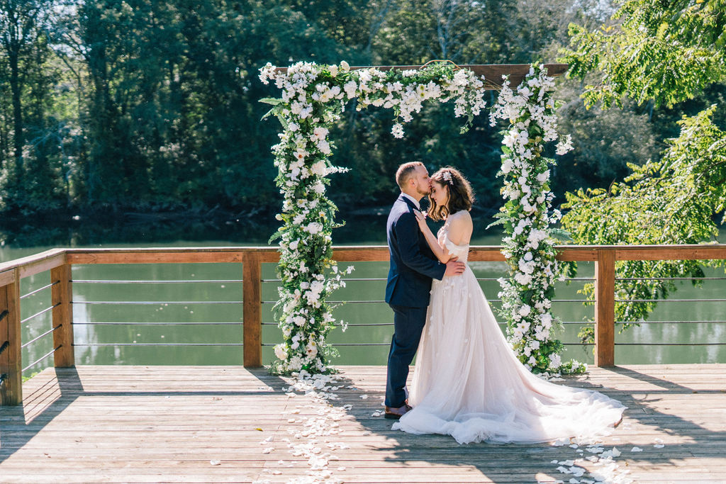 groom holding his brides waist and kissing her in front of their river side wedding under a flower and greenery arch.