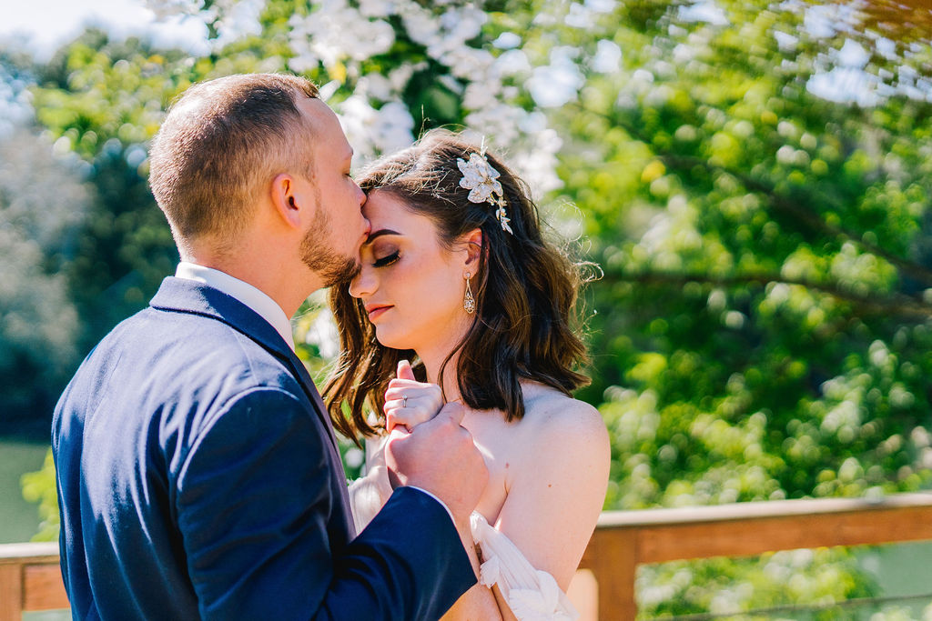 groom kissing his brides forehead right before they have their wedding, holding her hand with green trees in the background