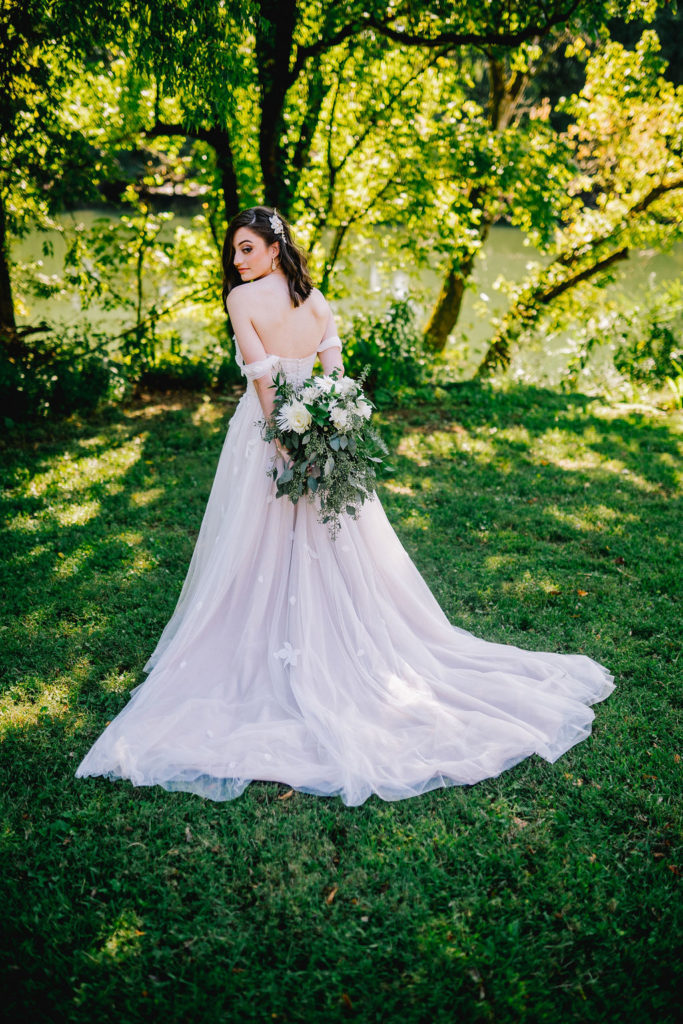 Bride in her wedding dress with the train spread out across the grass. Bride is holding her bouquet behind her back and looking over her shoulder