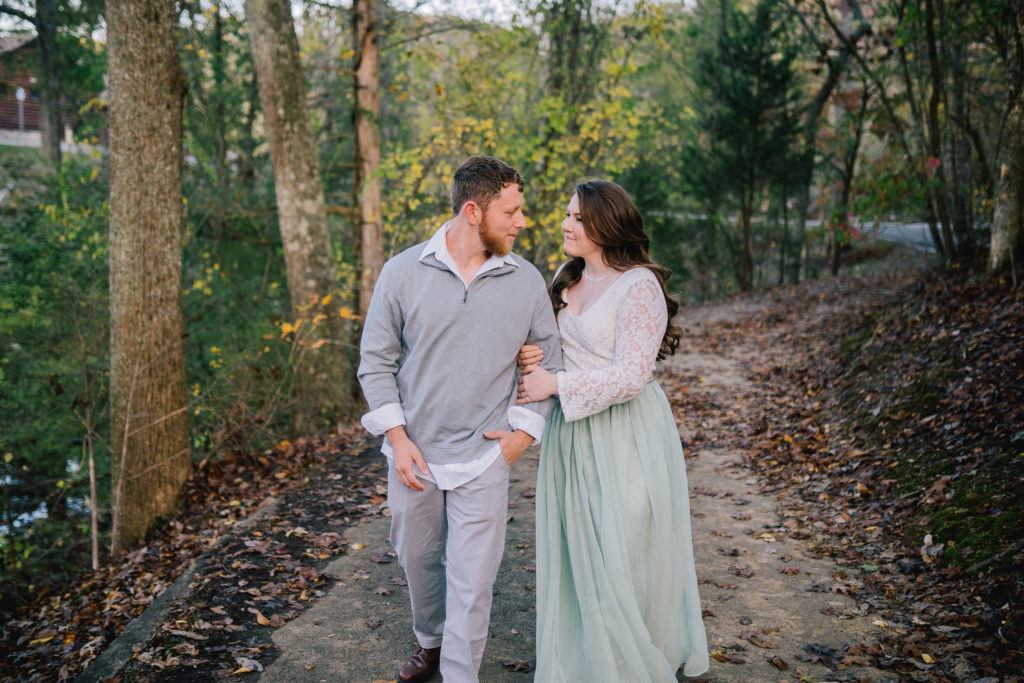 couple walking on a trail in the fall. Woman is holding the mans arm and they are looking at each other. Engagement session in Knoxville Tennessee