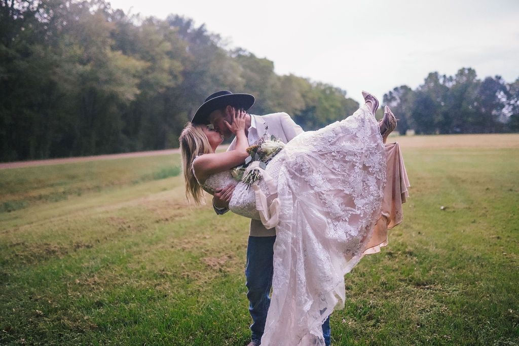 country couple. Groom is carrying bride in a Louisiana field. Bride is wearing a gorgeous lace mermaid gown celebrating and laughing after budget friendly wedding day