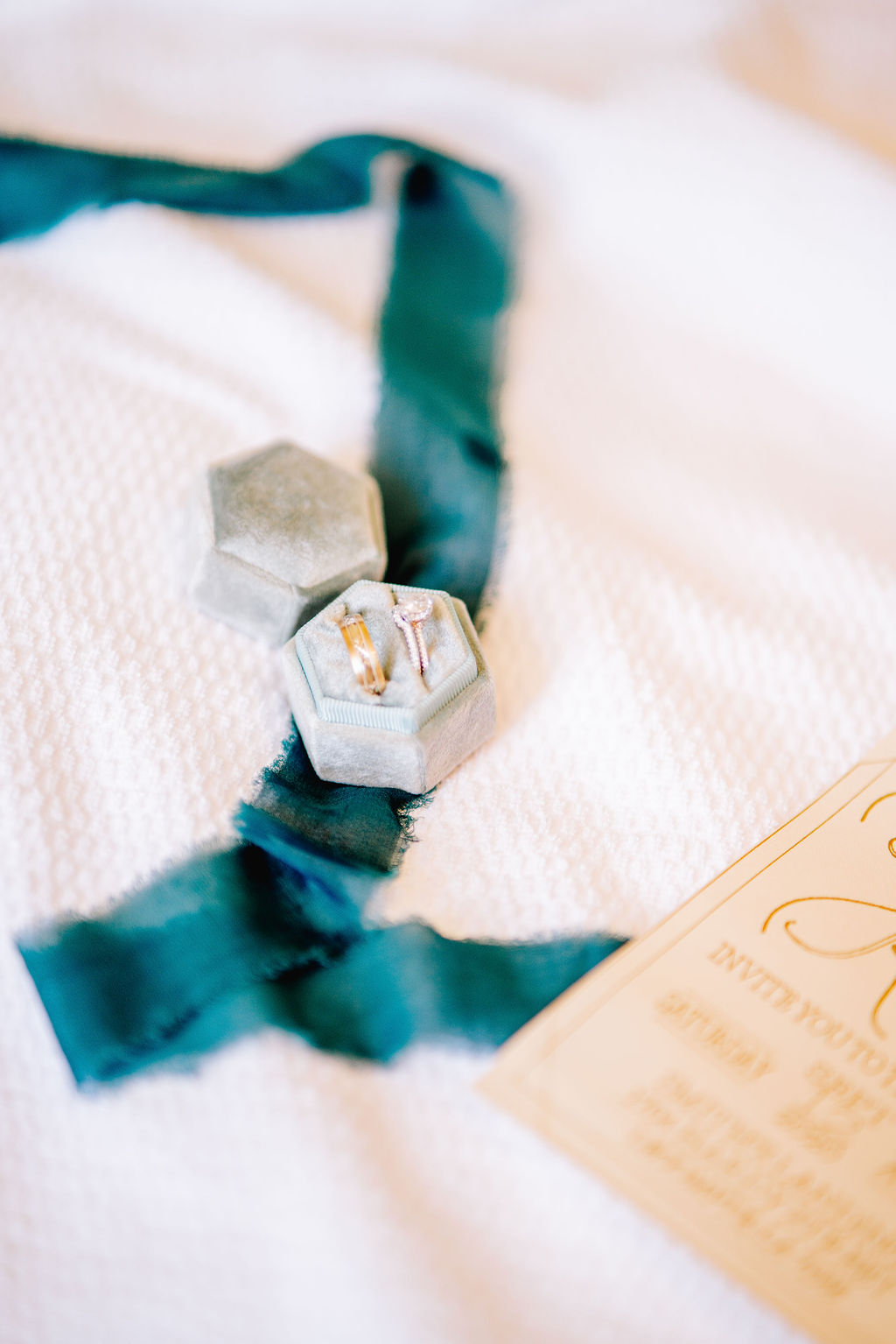 flat lay photo of wedding ring in box and blue ribbon with an invite to the wedding