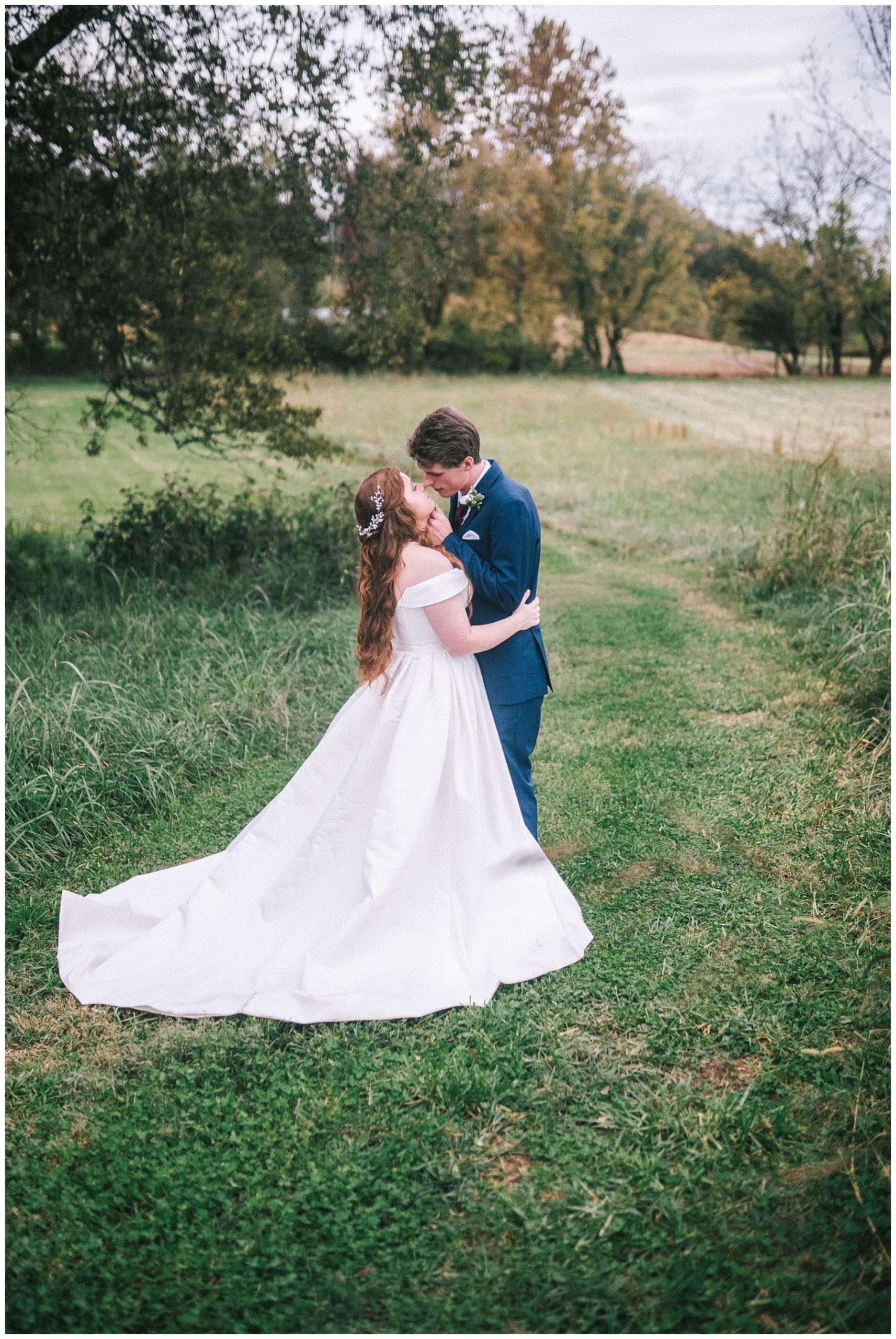 bride and groom standing in a field with trees kissing with the bride in a satin white gown that is off the shoulder and groom in a blue suit