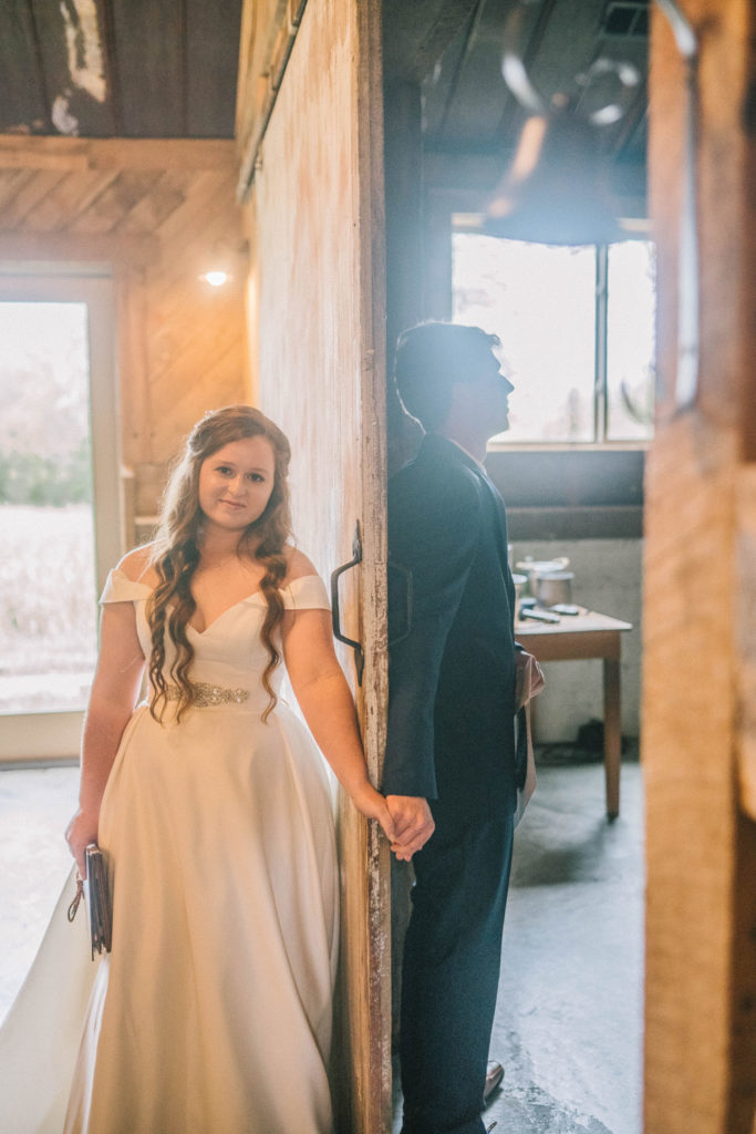 bride and groom standing on opposite sides of a barn door and holding hands before their first look at butler Farms