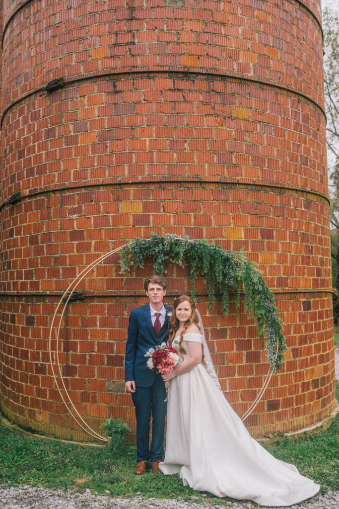 bride and groom standing infront of a brick wall under a gold circle arch with eucalyptus leaves hanging from the top. Very modern and rustic bridals.