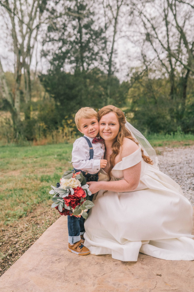 bride and little boy posing together in front of green trees on wedding day. bride is crouched down next to the little boy
