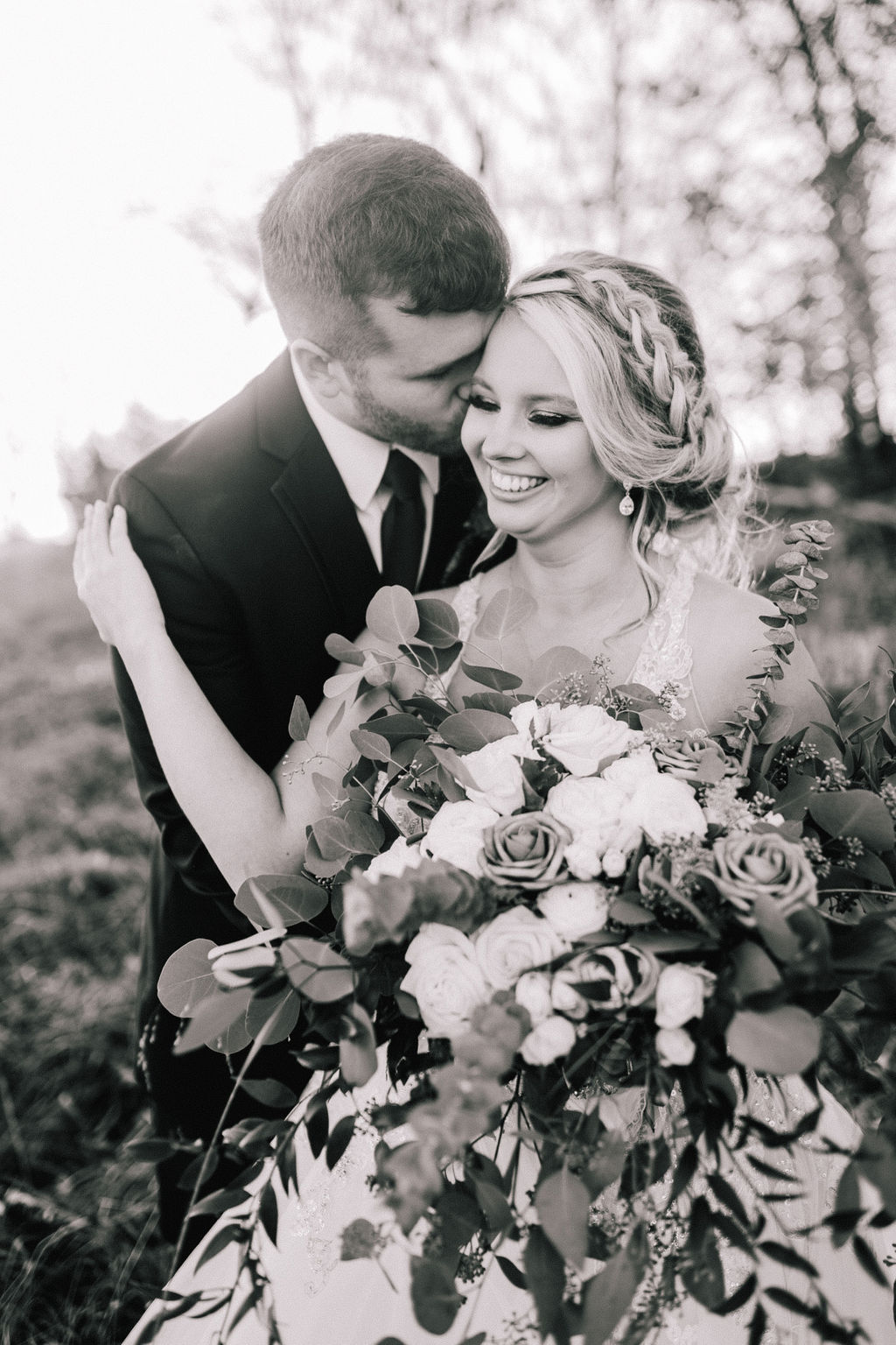 black and white image of bride and groom in a field for their destination wedding with the groom standing behind the bride as he kisses her cheek and the bride smiles and reaches back for him