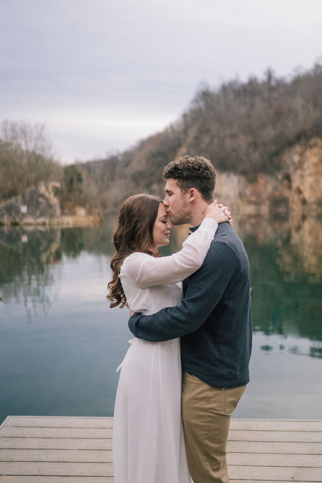 couple outside by the water holding each other as he kisses her forhead