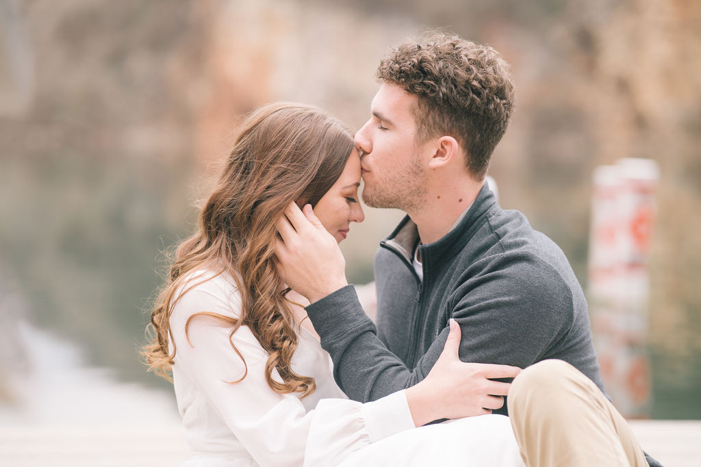 couple sitting down outside by a body of water while he kisses her forehead