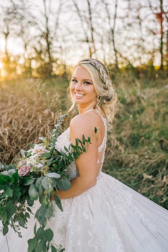 beautiful blonde bride in a floral lace ballgown dress poses in a field at sundown with her wedding bouquet