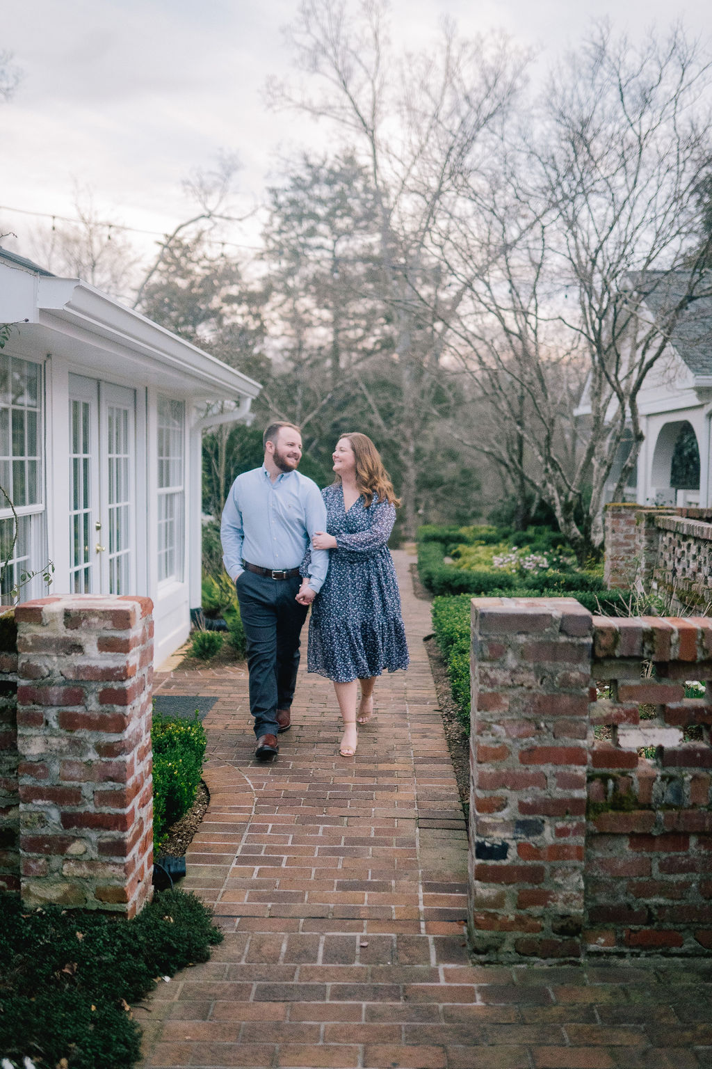 engagement session in the garden of a Knoxville home. Couple walking towards the camera past a greenhouse. Brick wall and stone path