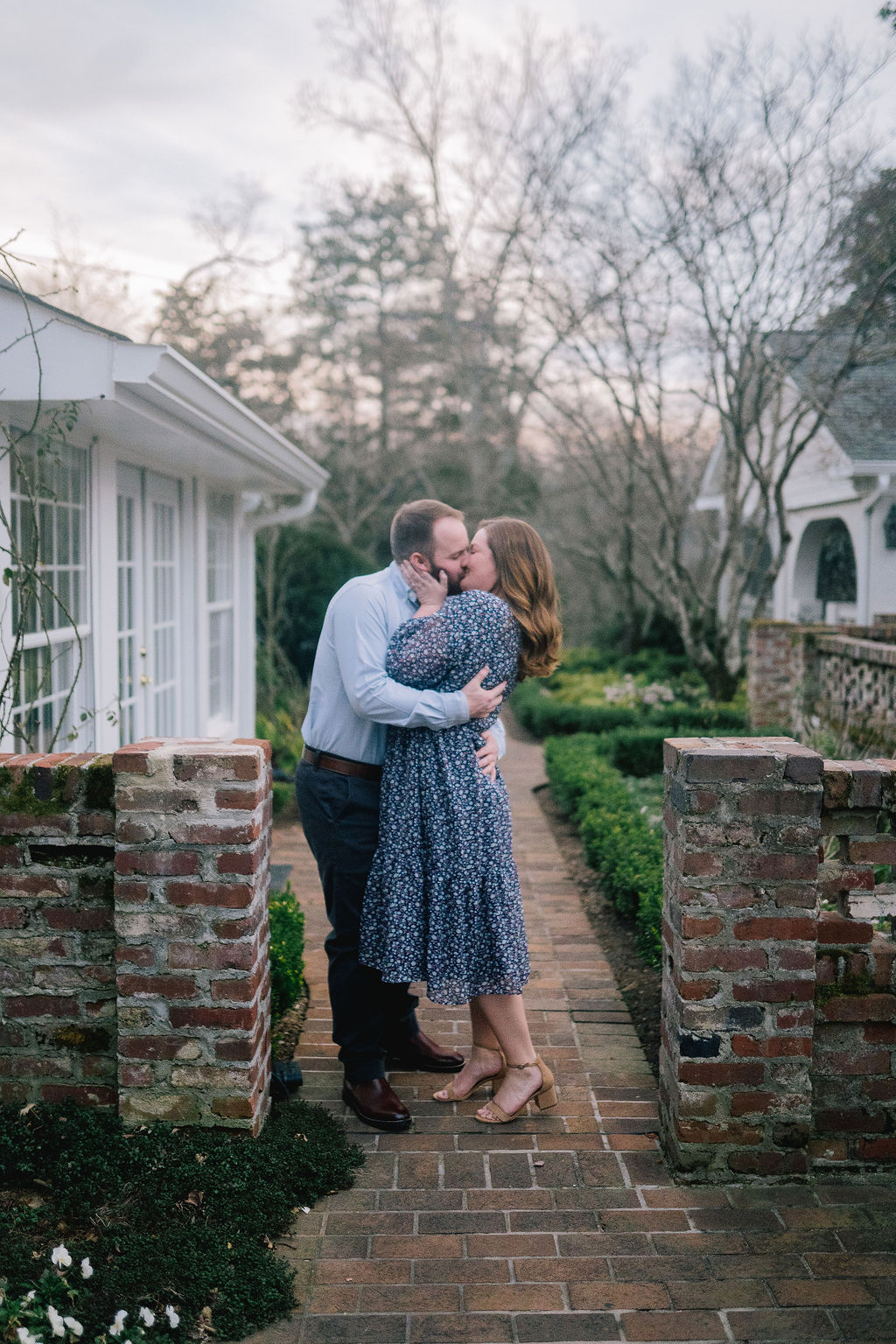 engagement session in the garden of a Knoxville home. Couple walking towards the camera past a greenhouse. Couple kissing at the opening of the brick wall.