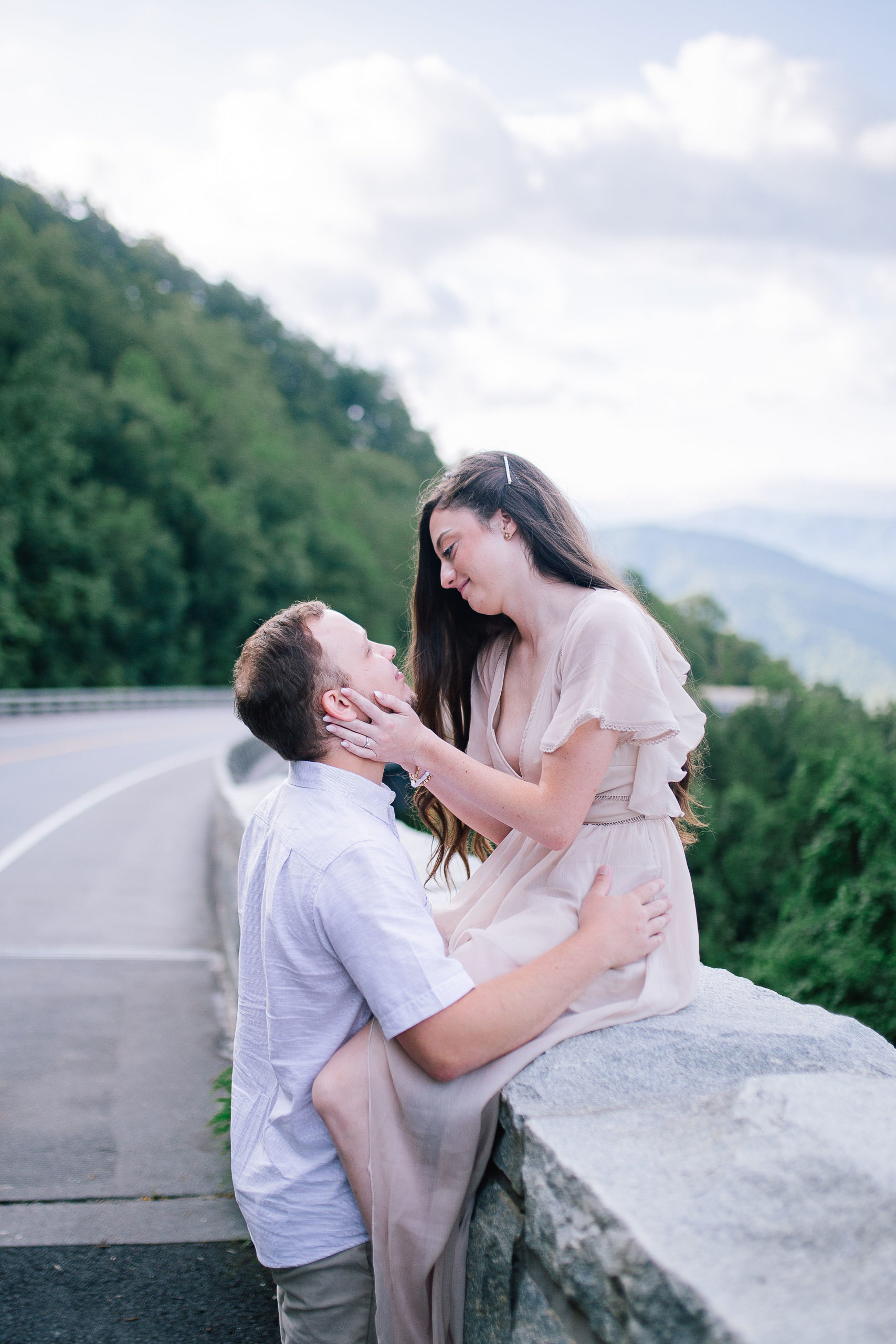smoky mountain foothills adventure session for engagements. Bride sitting on a stone wall while her groom holds her legs and they look out on the Foothills