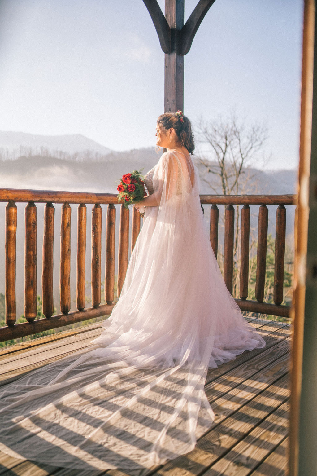 smoky mountain bride on her wedding day surrounded by fog as she looks out the balcony wearing her sparkly wedding gown and holding her red roses