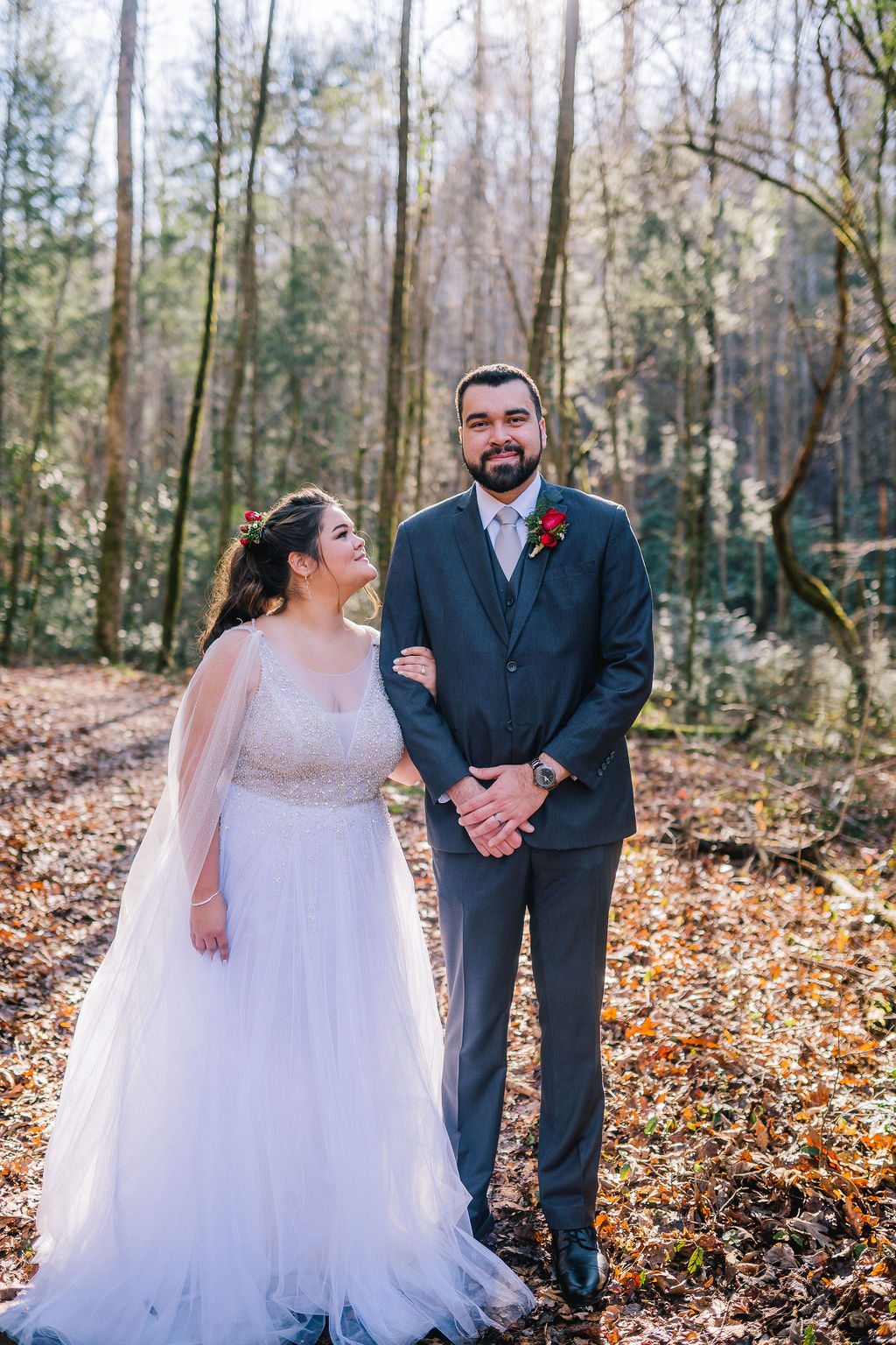 bride and groom standing together on their wedding day in the smokies in the forest. grrom is looking and smiling at ther camera, while his bride looks up and smiles at him