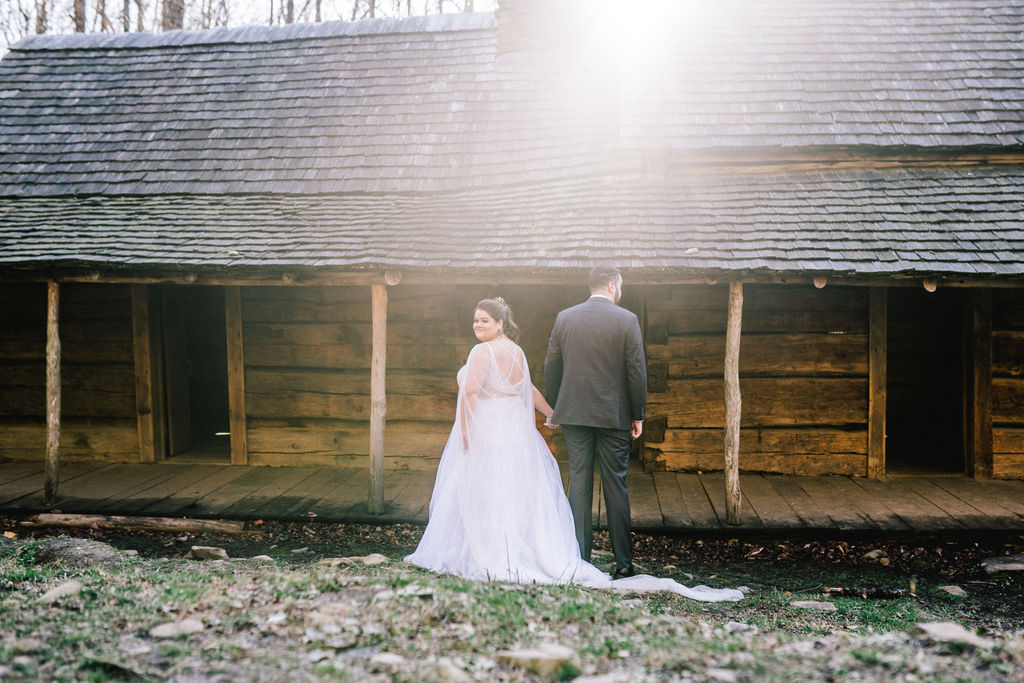 romantic wedding day in the smokies at a cabin in the woods. Bride and groom walking towards to cabin, sun flares over the roof.