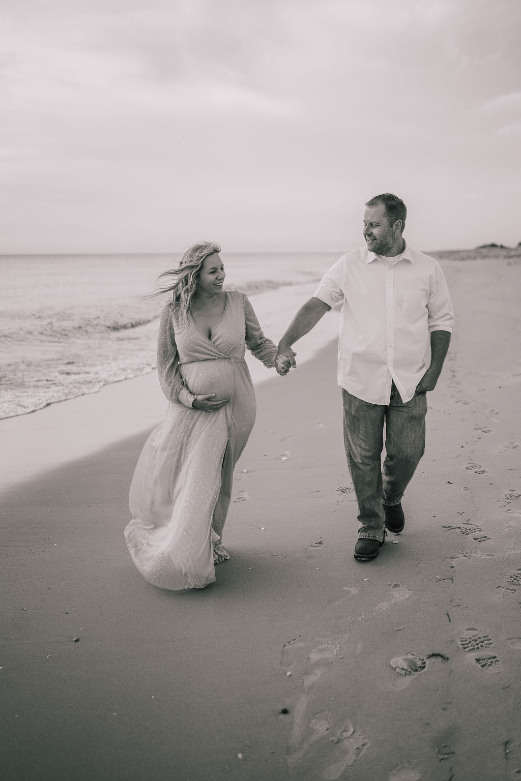 black and white photo of a couple walking on the shore line of North Carolina's coast for their dreamy maternity session. Woman is pregnant and holding her bump with one hands and holding her husbands with the other