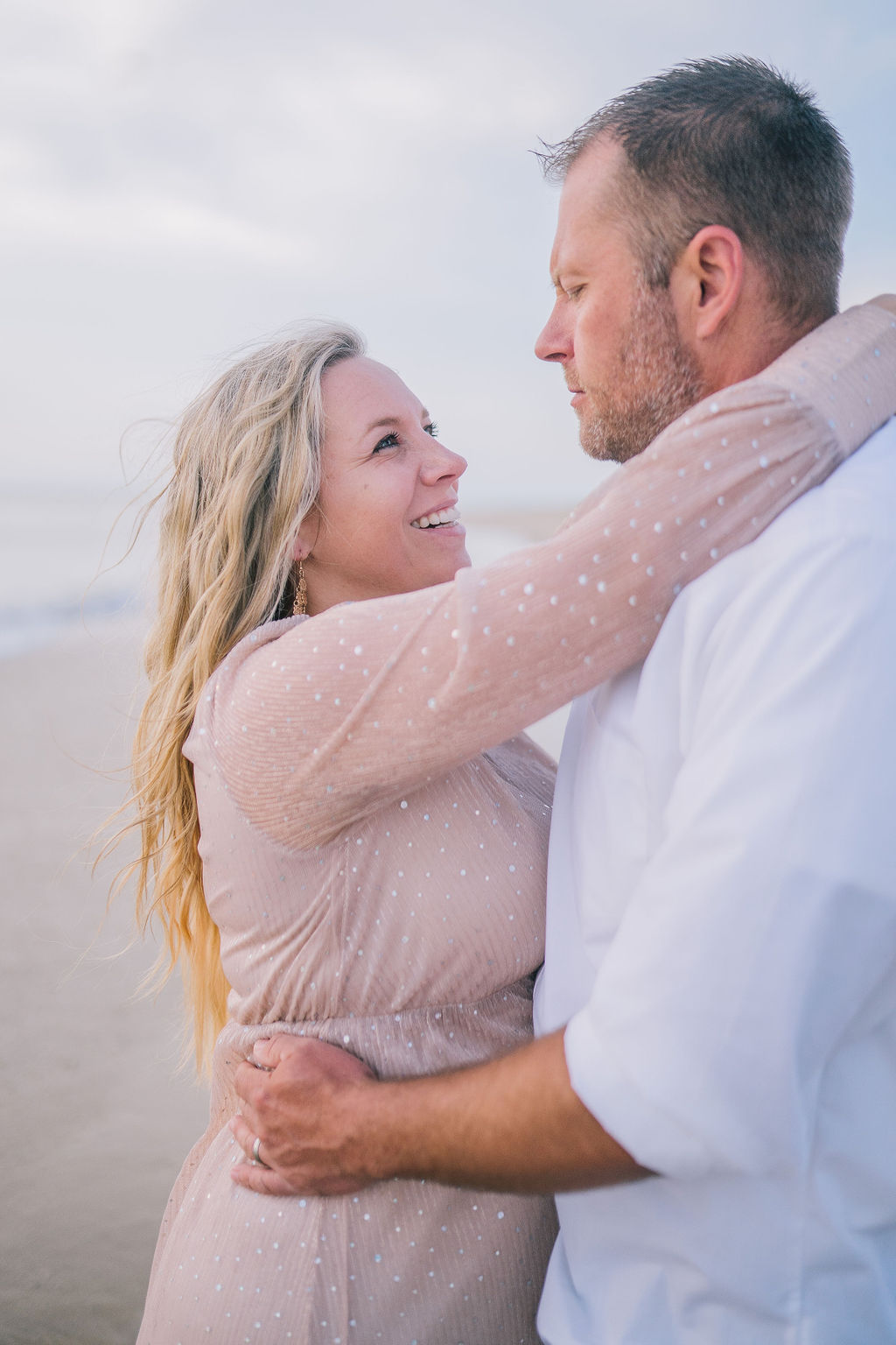 woman has her arms around mans neck, smiling at him as he embraces her waist. Maternity session at the Outerbanks.