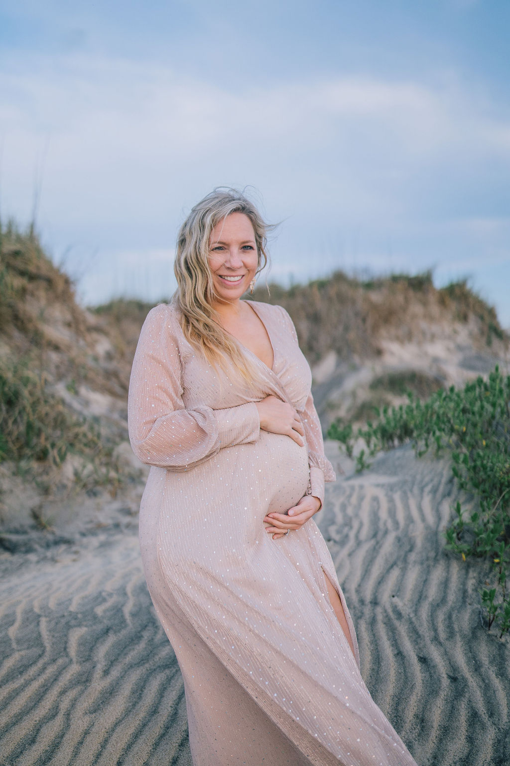Outerbanks Maternity Session with woman standing in the sand with hills of beach grass behind her as she holds her bump and the wind blows through her hair.