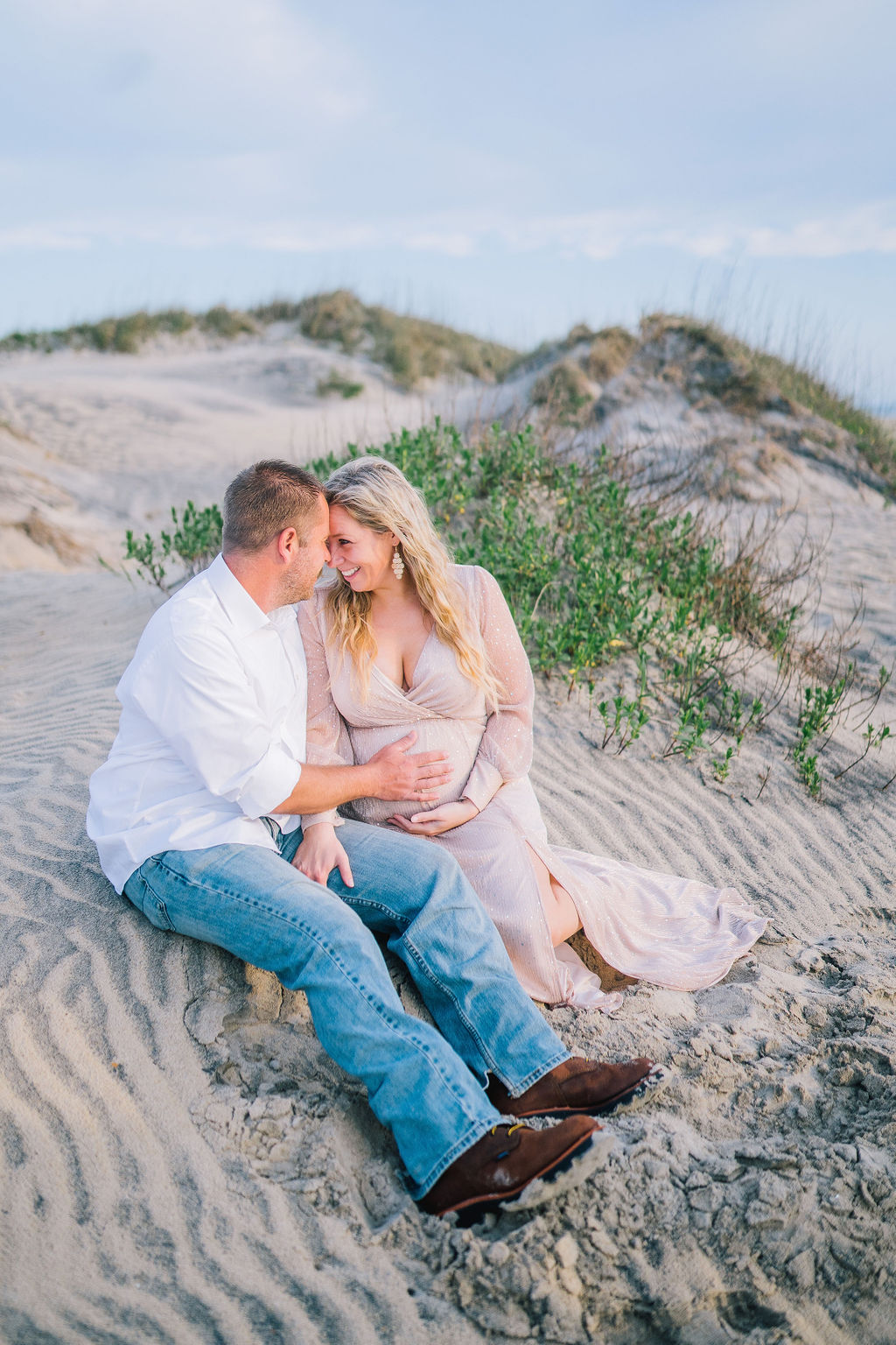 husband and wife are pregant at the beach in Outerbanks for their maternity session. They are sitting together on the sand with tall grass in the background
