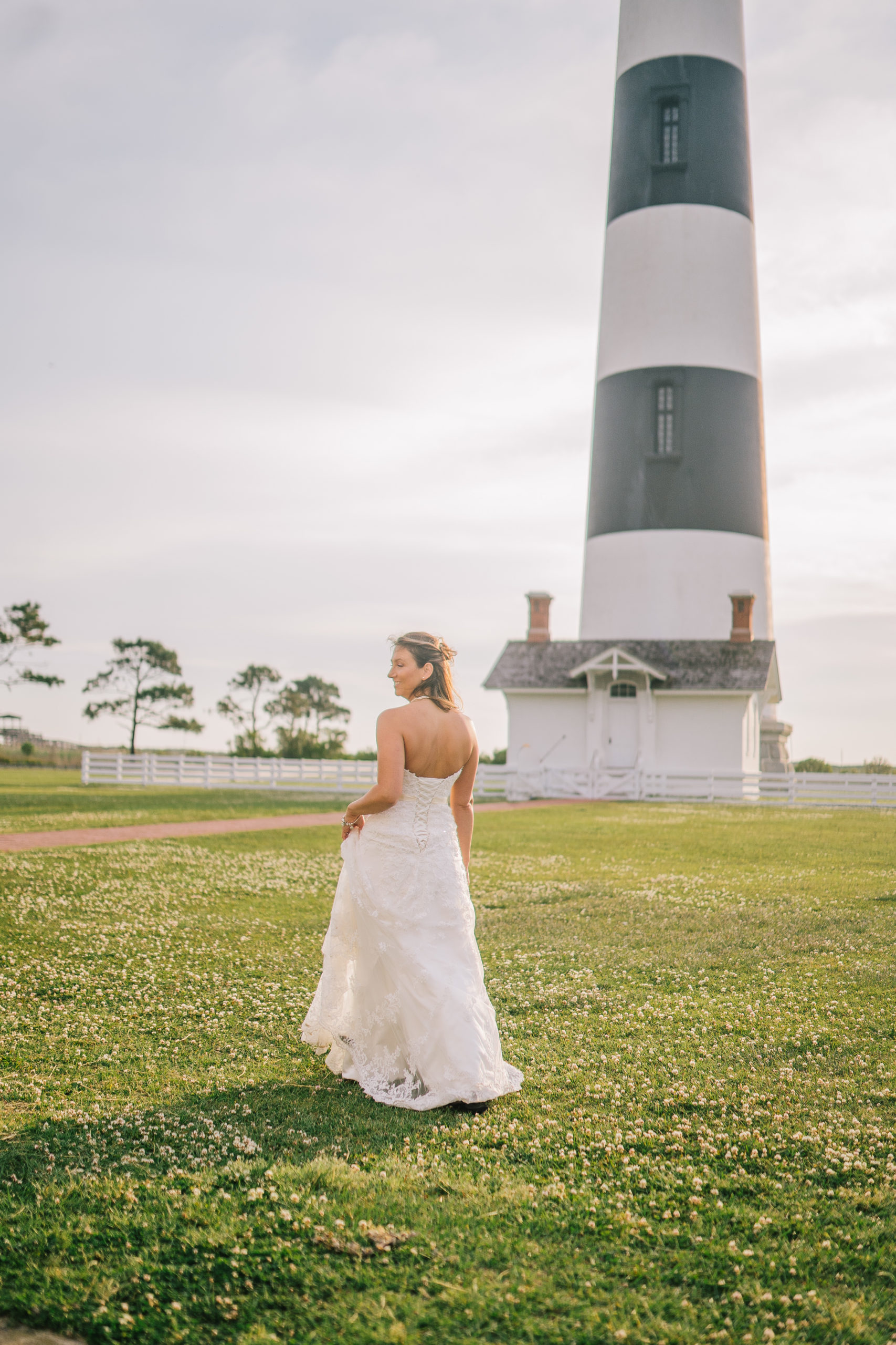 Bride ina lave mermaid gown walks on a green field on the East coast towards a striped black and white light house