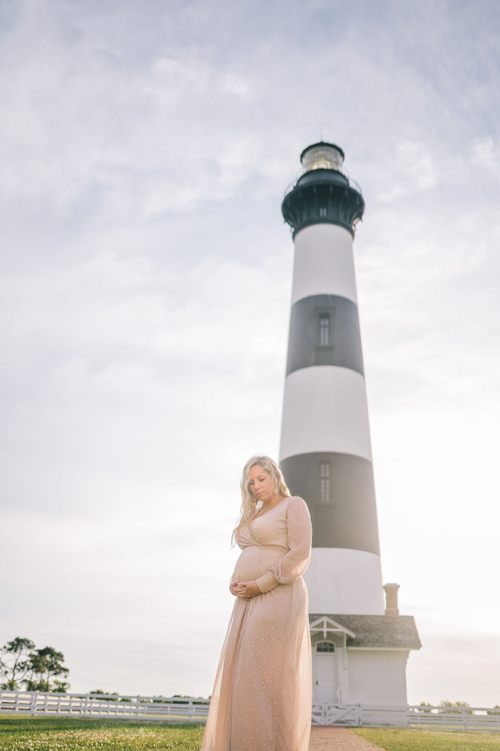 lighthouse maternity session in the outerbanks, the pregnant woman is wearing a blush pink formal gown and is holding her bump in front of a black and white striped lighthouse